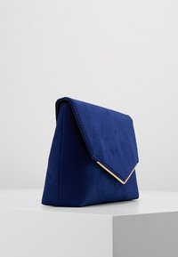 Bolso de mano de ante azul marino con una solapa triangular y herrajes en tono dorado. Superficie texturizada, forma angular y diseño minimalista.