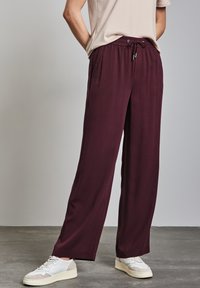 Pantalon large en bourgogne, léger, avec une taille élastique et un cordon de serrage, associé à des baskets blanches sur une surface grise texturée.