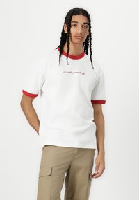 White short-sleeve T-shirt with red ringer collar and sleeves. Features embroidered text on the front. Worn with light khaki cargo pants.