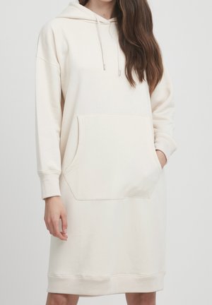 Vrouw draagt een lange, crème kleurige hoodiejurk met voorzak en trekkoorden, staand tegen een effen lichte achtergrond.