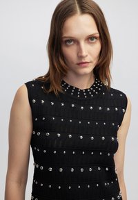 Abito nero senza maniche con un design a maglia testurizzata, caratterizzato da borchie metalliche disposte in file orizzontali e una scollatura alta.