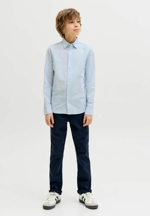 Dječak u svijetloplavoj košulji s gumbima, tamnoplavim hlačama i bijelim tenisicama stoji i gleda blago u stranu na jednobojnoj pozadini.
