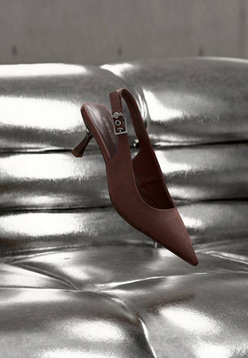 Καφέ μυτερό παπούτσι slingback με χαμηλό τακούνι και λουράκι με πόρπη που ακουμπά σε επιφάνεια από γυαλιστερό, μεταλλικό ασημί ύφασμα με καπιτονέ.