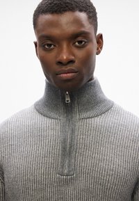 Suéter gris de canalé con un cuello alto y cremallera frontal. La tela tiene una apariencia texturada con un brillo sutil y un acabado suave.