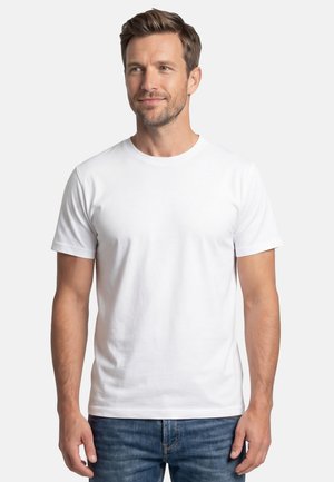 Mann mit kurzen braunen Haaren, der ein einfaches weißes T-Shirt mit kurzen Ärmeln und blaue Jeans trägt, vor einem weißen Hintergrund stehend.