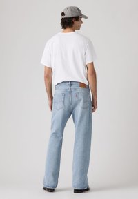 Άνδρας που φορά γκρι καπέλο, λευκό t-shirt και ανοιχτό μπλε τζιν με ίσια γραμμή, στέκεται με την πλάτη στην κάμερα σε ουδέτερο φόντο.