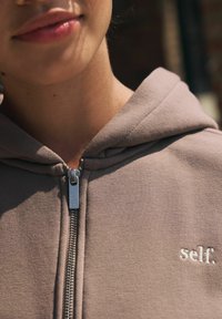 Prindi särk tummas pruunis kangas, millel on hall tõmblukk ja valge tikandiga "self." logo rinnal. Pehme tekstuur.