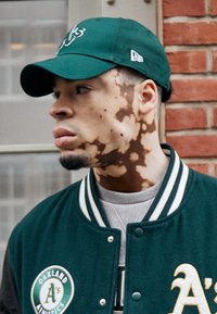 Πράσινο καπέλο μπέιζμπολ και πράσινο παρκά με λευκές ρίγες, με το λογότυπο των Oakland Athletics, φορεμένο πάνω από έναν γκρίζο πουλόβερ.