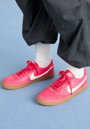 Pies con zapatillas Nike rosa brillante con logotipos de swoosh blancos, calcetines blancos y pantalones negros holgados sobre un suelo azul.