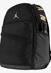 Rucsac din țesătură neagră, cu un logo Jumpman auriu, un compartiment principal cu fermoar și o buzunar lateral pentru o sticlă de apă.
