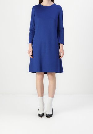 Mujer con un vestido azul de manga larga hasta la rodilla, calcetines blancos y zapatos negros de punta, posando frente a un fondo blanco liso.