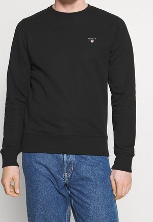Homme portant un pull noir à manches longues et col rond avec un petit logo GANT sur la poitrine, associé à un jean en denim bleu.