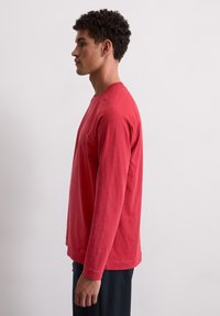 Rode longsleeve shirt van zacht materiaal, met een ronde hals en een relaxte pasvorm. Draagt met donkere broek, getoond in profiel.