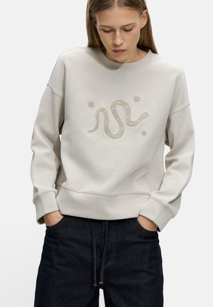Bluza w jasnym beżowym kolorze, wykonana z bawełny, z długimi rękawami, ściągaczami i wyszywanym wzorem węża w stonowanej kolorystyce na środku.