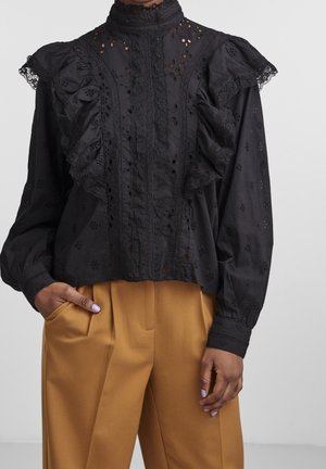 Femme portant un chemisier en dentelle noire avec des volants et un col montant, associé à un pantalon large couleur moutarde, main dans la poche, ongles violets clairs visibles.