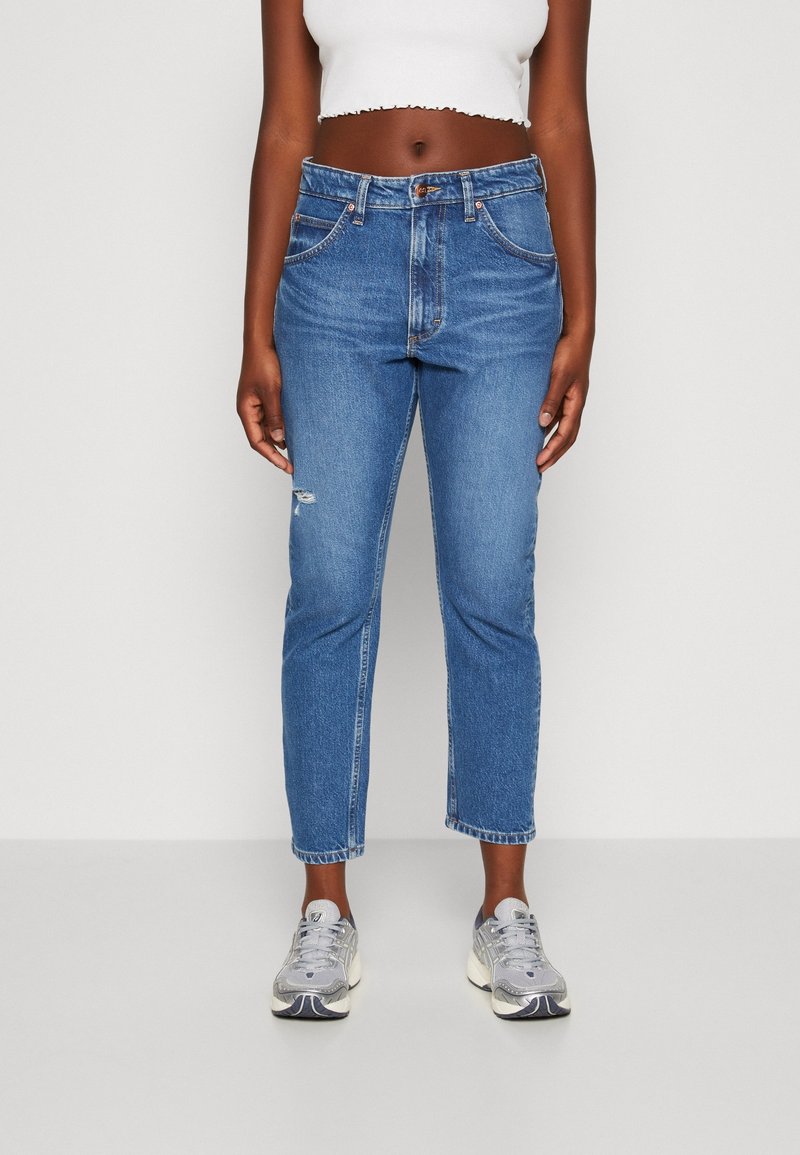 Jean en denim bleu avec une taille haute et une coupe légèrement raccourcie. Présente des détails usés au genou gauche. Porté avec des baskets grises.