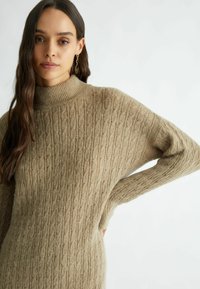 Maglione beige lavorato a maglia con colletto alto a coste, caratterizzato da un motivo texturizzato e una vestibilità ampia. Il tessuto appare morbido e caldo.