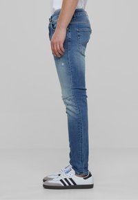 Kék farmerből készült skinny farmernadrág, enyhén elhalványult felülettel, apró kopásokkal és sima textúrával. Fehér és szürke tornacipővel párosítva.