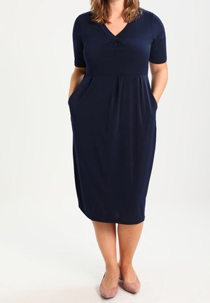Mujer con vestido azul marino hasta la rodilla con mangas cortas y bolsillos, combinado con zapatos planos rosas claros, de pie sobre un fondo blanco.