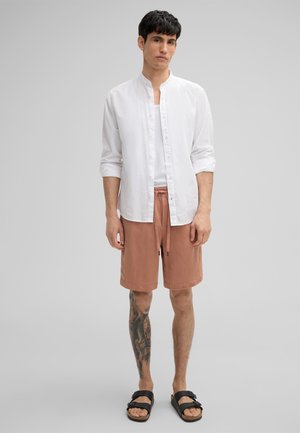 Weißes Leinen-Hemd mit hohen Kragen, kombiniert mit pfirsichfarbenen Shorts mit elastischem Bund und schwarzen Sandalen.