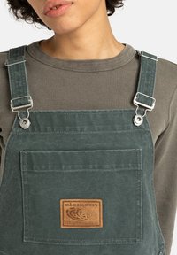 Salopette verde in tela con tasca frontale e etichetta in pelle. Le bretelle regolabili presentano hardware argentato; indossata sopra una maglietta grigia a coste.