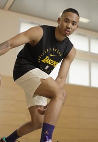 Camisola preta sem mangas com o logo amarelo dos "Lakers", calções claros e meias roxas. Ele está a driblar uma bola de basquetebol num ginásio.