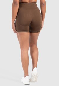 Shorts marroni a vita alta realizzati in tessuto elastico, con una texture liscia e una lunghezza fino al ginocchio, abbinati a scarpe da ginnastica bianche.