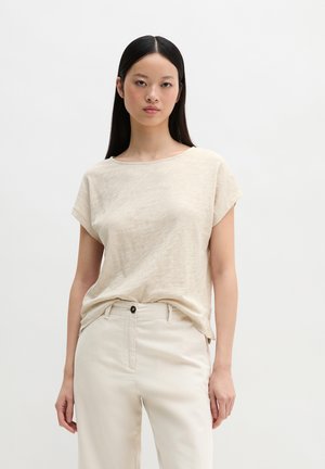 Vrouw met lang donker haar, gekleed in een beige top met korte mouwen en crèmekleurige, hooggetailleerde broek, staand tegen een effen witte achtergrond.
