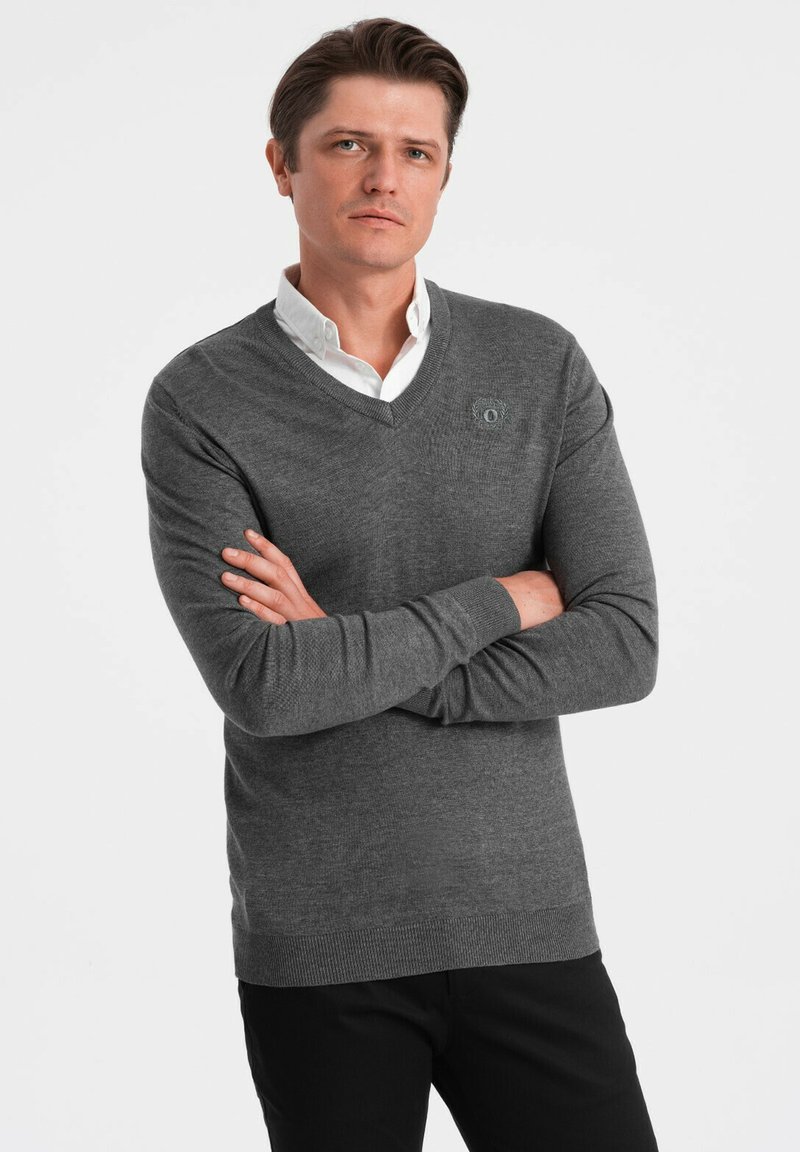 Pull gris avec un col en V, fabriqué en matière douce. Doté d'un logo subtil sur la poitrine et de poignets et un ourlet côtelés. Porté par-dessus une chemise blanche.