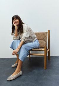Flaturi bej țesute cu vârful ascuțit, așezate pe un podea închisă la culoare. Modelul poartă o fustă albastră deschisă și o jachetă crem, stând pe un scaun din lemn.