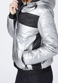 Silver- och svart puffjacka med huva, som har en blandning av glänsande och matta texturer, dragkedjefickor och kontrasterande paneldesign.