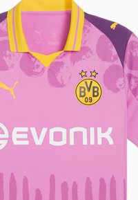 Camiseta de fútbol rosa con un hombro púrpura, cuello amarillo y logotipo negro. Presenta patrones abstractos y texto de patrocinador en blanco.