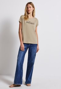 Camiseta de algodón beige con la palabra "reflect" impresa en marrón, combinada con unos vaqueros de denim azul holgados y sandalias planas color marrón claro.