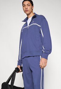 Βασιλικό μπλε φόρμα με λευκές ρίγες στα πλαϊνά, ζακέτα με φερμουάρ και ταιριαστό παντελόνι. Κατασκευασμένη από μαλακό ύφασμα, με εφαρμοστή σχεδίαση και τσέπες.