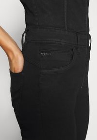 Gros plan sur une personne portant un pantalon en denim noir G-Star, avec la main dans la poche arrière, sur un fond uni clair.