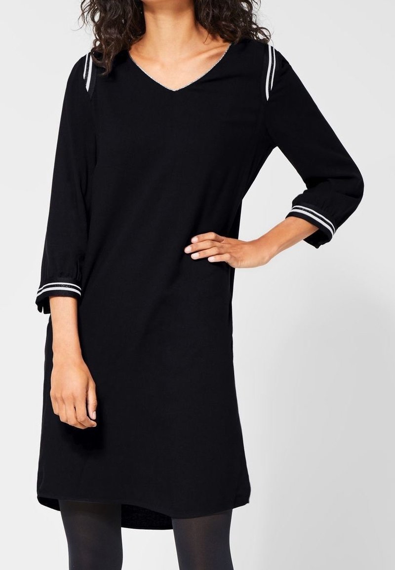 Femme portant une robe noire longueur genou avec manches trois-quarts et bordure rayée blanche aux épaules et aux poignets.