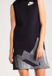 Svart ärmlös klänning med Nike-logotyp på bröstet; har ett strukturerat grått geometriskt mönster vid fållen. Tillverkad av mjukt, elastiskt material.