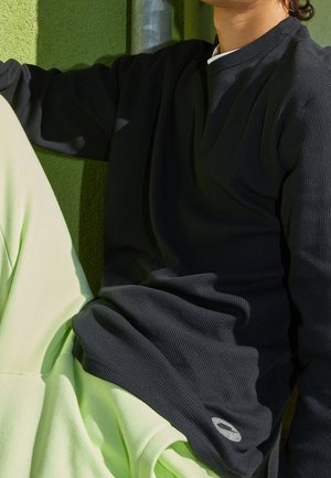 Schwarzes, geripptes Langarmshirt mit rundem Ausschnitt, kombiniert mit hellgrünen Jogginghosen. Verfügt über ein kleines rundes Logo am Saum.