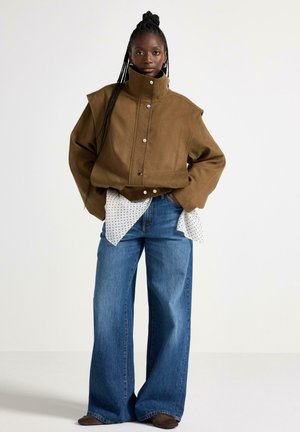 Giacca marrone oversize con colletto alto, dotata di bottoni a pressione. Sotto, una camicia bianca a pois. I pantaloni larghi in denim blu completano l'outfit.