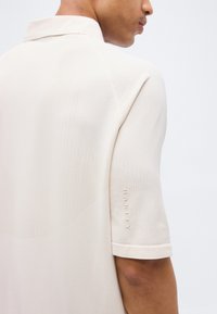 Άνδρας που φοράει κοντομάνικο ριμπ πικέ πουκάμισο πόλο σε εκρού χρώμα με τη λέξη "Oakley" κεντημένη στο μανίκι, εμφανίζεται από πίσω και πλάγια.