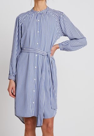 Kvinde iført en blå og hvid vertikalt striped skjortekjole med lange ærmer og et matchende bælte bundet i taljen.