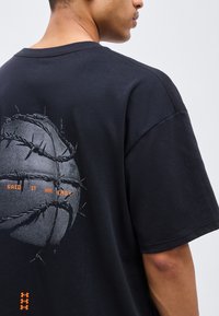 Άνδρας που φοράει μαύρο μπλουζάκι με μπάλα του μπάσκετ τυλιγμένη με συρματοπλέγματα και κείμενο «ΕΙΠΑ ΟΤΙ ΗΤΑΝ ΕΥΚΟΛΟ» στην πλάτη.