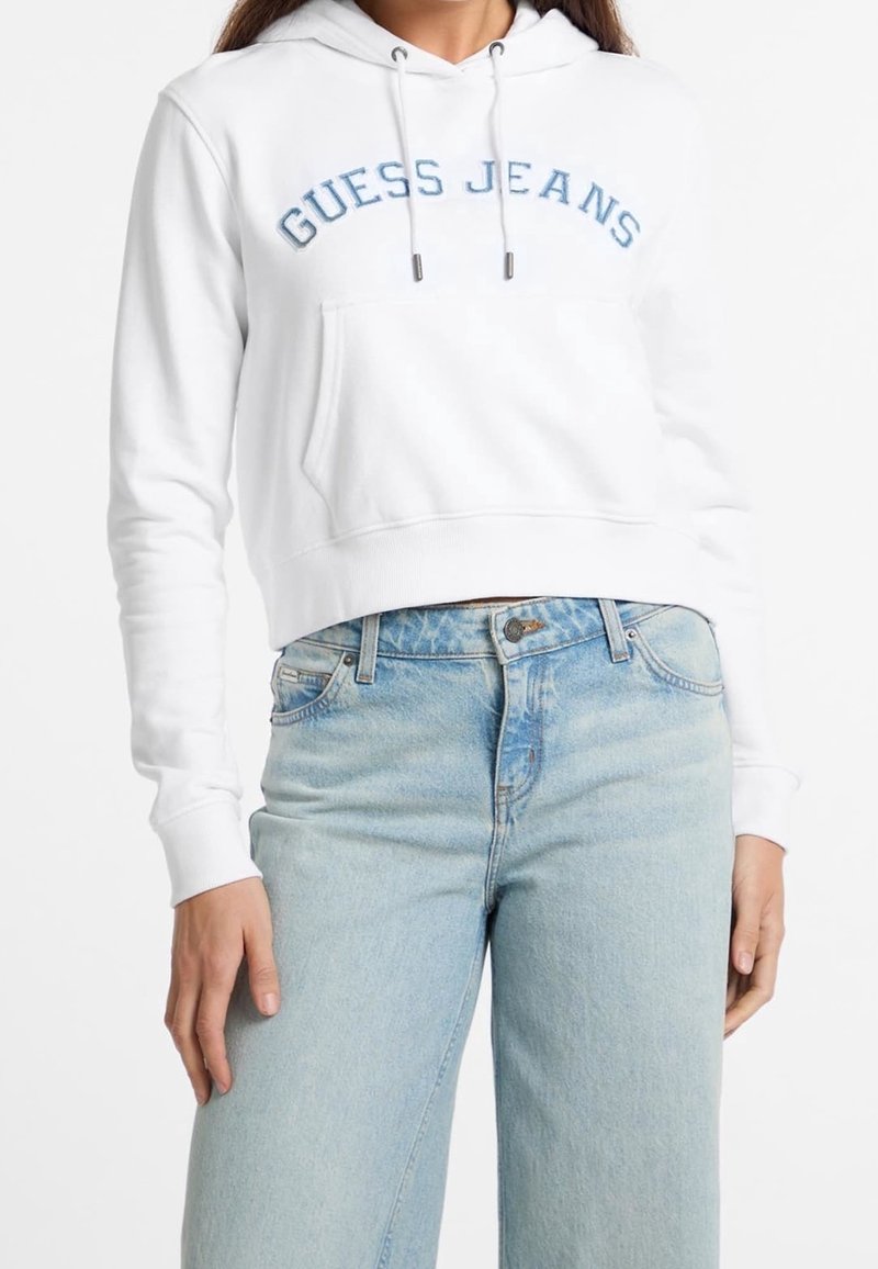 Femme portant un sweat à capuche court blanc avec le texte bleu "Guess Jeans" et un jean denim taille haute bleu clair, debout bras détendus.