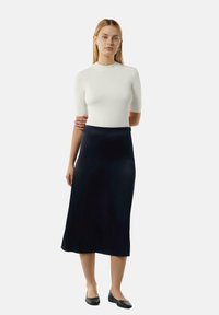 Weißes, tailliertes Oberteil mit kurzen Ärmeln und hohem Ausschnitt, kombiniert mit einem langen, marineblauen, seidenen Rock. Schwarze flache Schuhe runden den Look ab.