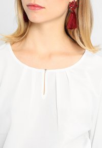 Weiße Bluse mit Schlüsselloch-Ausschnitt, gefälteltem Detail und glatter Textur. Kombiniert mit roten Quastenohrringen mit floralen Akzenten.