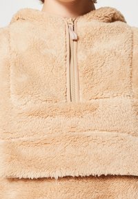 Pullover in pile beige con chiusura a mezza zip, caratterizzato da una texture morbida e un cappuccio. Il design include una grande tasca frontale.