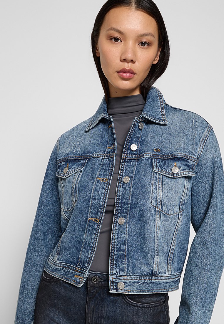 Chaqueta de mezclilla azul con un diseño corto, dos bolsillos en el pecho, botones plateados y costuras en contraste. Textura suave.