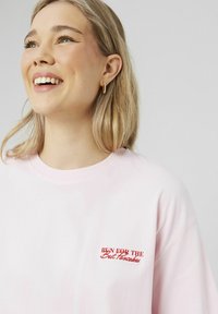 T-shirt en coton rose clair à col rond, présentant du texte brodé rouge qui dit "COURIR POUR LES MEILLEURES Crêpes." Texture lisse.