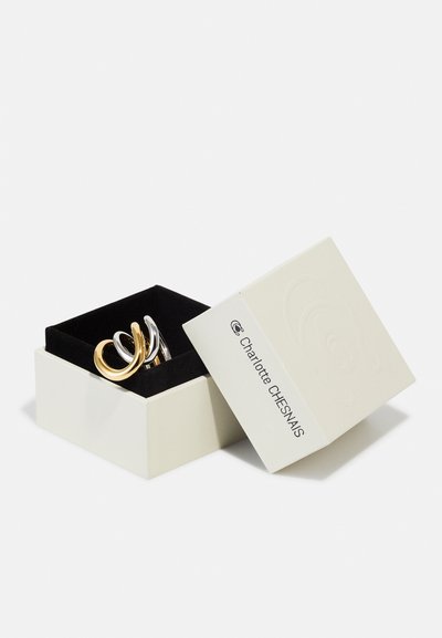 Avatud kreemjas ehetekast musta sisuga, mis hoiab kuldset ja hõbedast keerduv sõrmust, kaas on kaldu ja näitab Charlotte Chesnais logo.