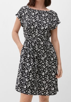 Vrouw draagt een korte mouwen jurk met zwart-witte dierenprint, met een strik in de taille en handen in de zakken, tegen een effen achtergrond.